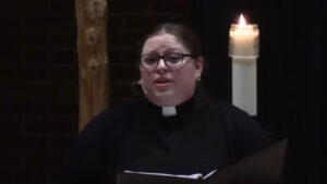 Angela LaMoore preaching in Nativity Church's sanctuary with a white lit candle behind her and a part of a wooden cross in the background